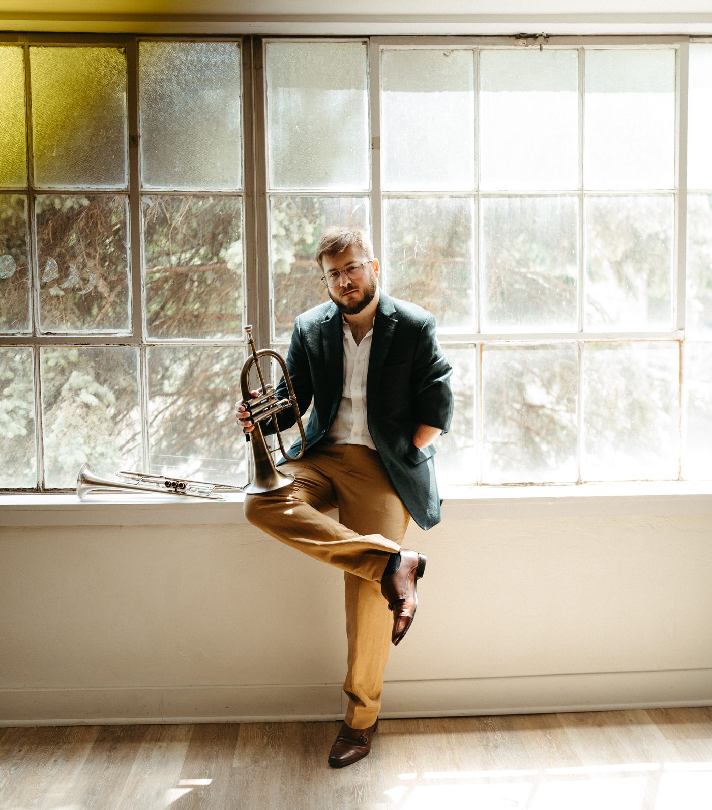 Christian Dyble seated on a sunlit windowsill in a dark green blazer, holding a flugelhorn, with a trumpet resting beside him